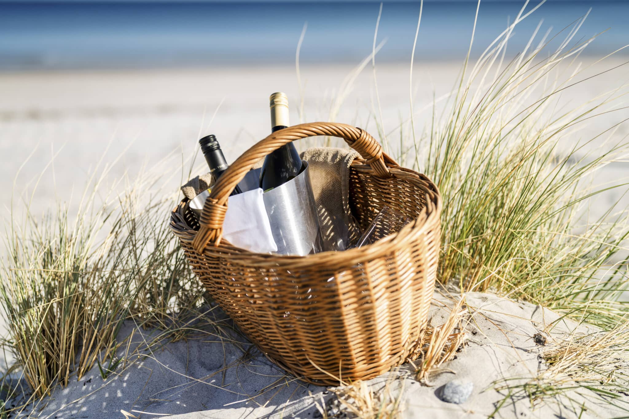 Weidenkorb mit Weinflaschen auf Sand, das Meer dahinter verschwommen.