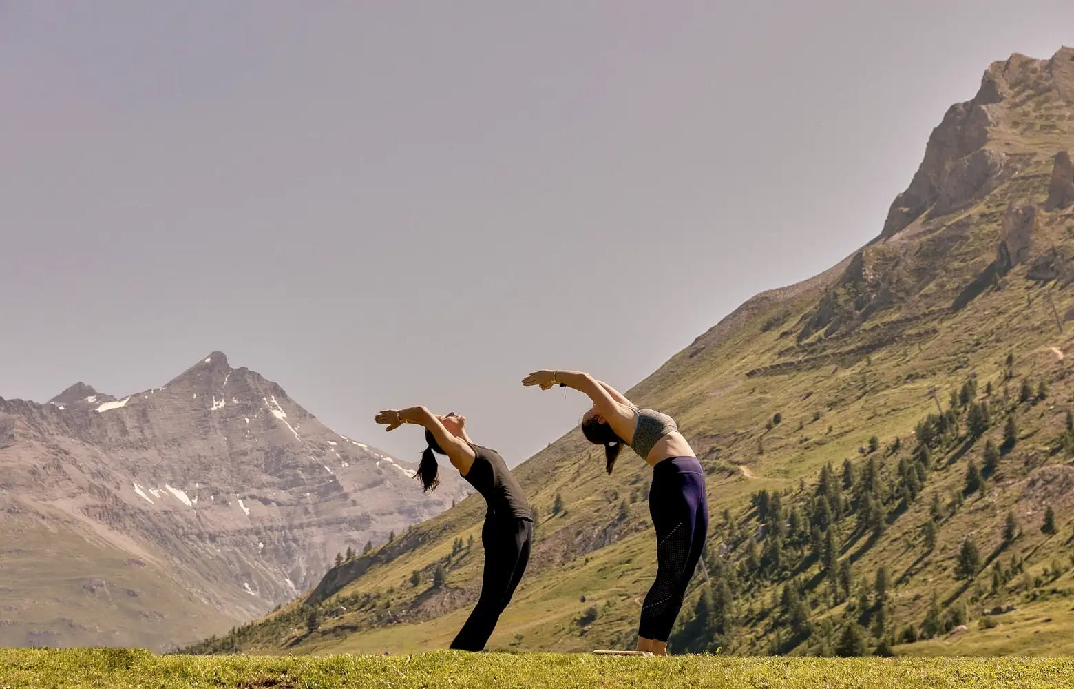 Zwei Menschen machen Yoga im Freien, dahinter Berge und klarer Himmel.