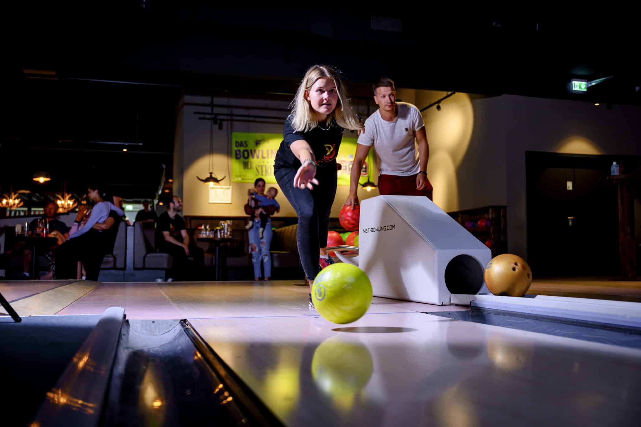 Eine Frau kegelt eine grüne Kugel, während andere Personen im Hintergrund zu sehen sind.