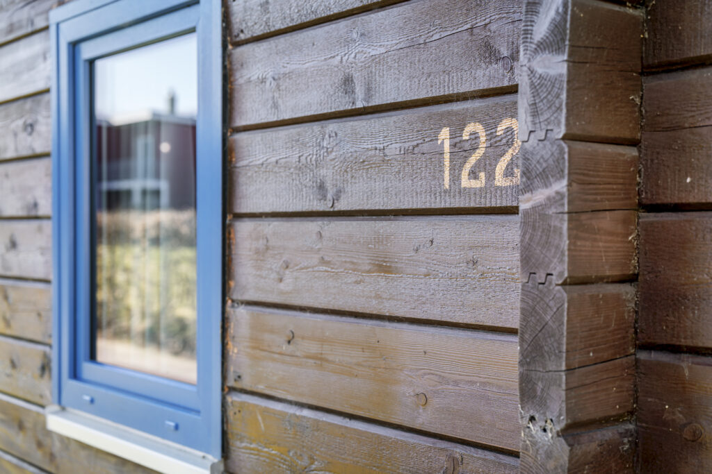 Nahaufnahme: braunes Holzhaus, blaues Fenster, weiße Nummer "122".