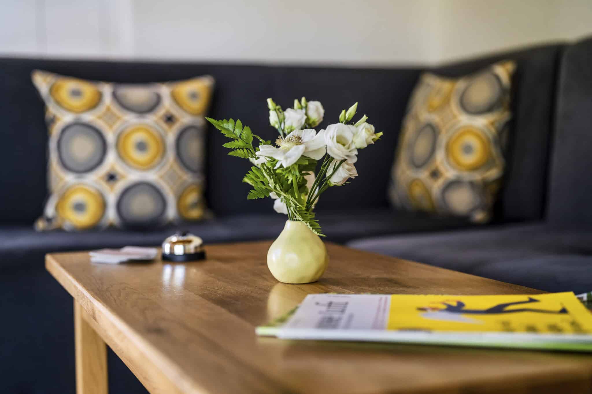Weiße Blumen in Vase, gelbe Zeitschrift, Glocke; dahinter Sofa.