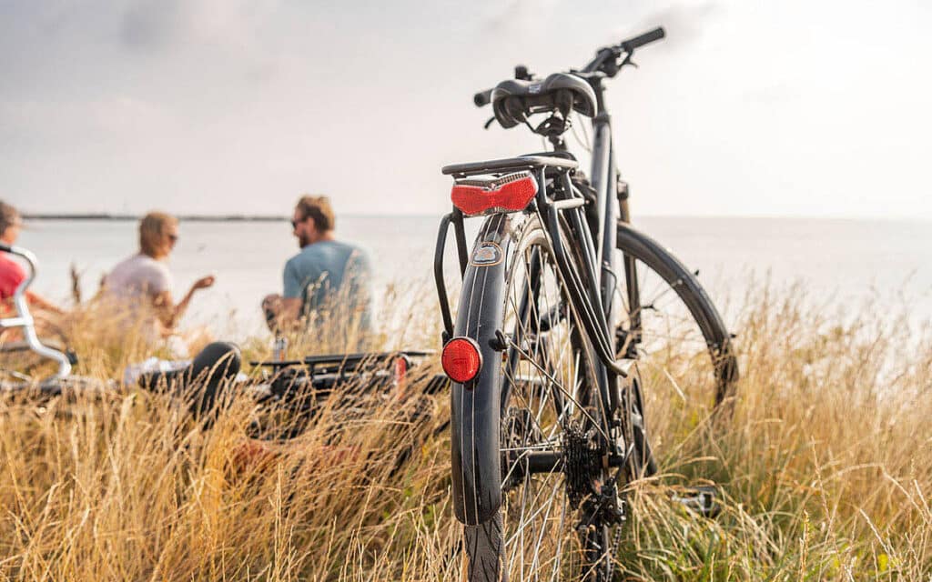 Fahrrad im Gras am Wasser; drei Personen entspannen sich vor dem Hotel.