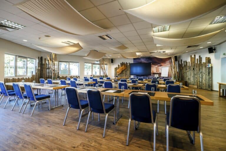 Heller Konferenzraum mit Tischen, blauen Stühlen, Bildschirm und Fenstern.