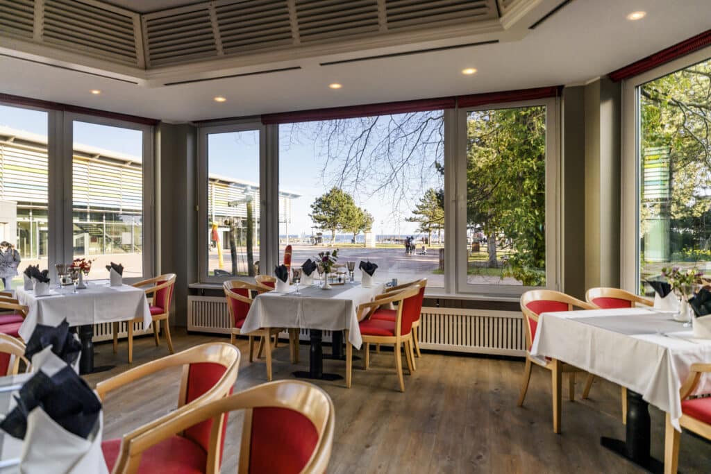 Sonniges Restaurant: gedeckte Tische, rote Stühle, Blick auf das Wasser und die Bäume.