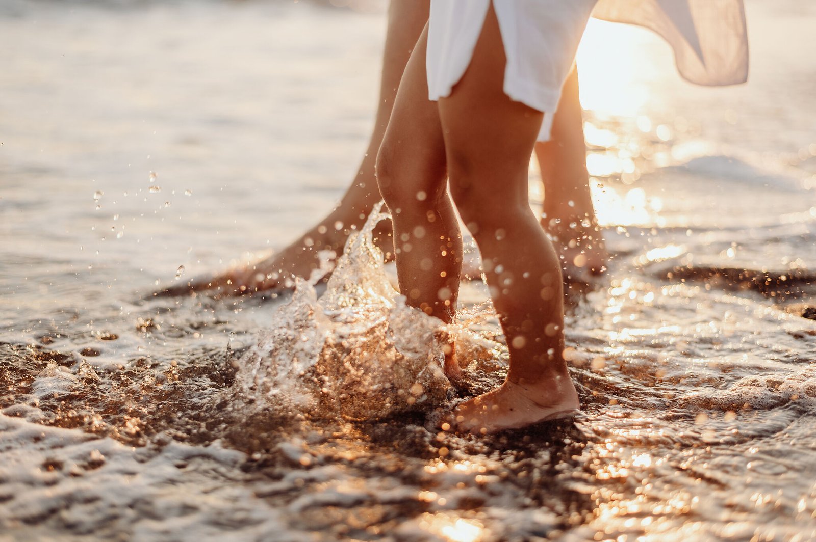 Kinderfüße plätschern im sonnenbeschienenen Strandwasser neben Erwachsenenbeinen.