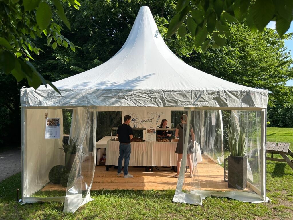 Weißes Zelt auf einer Wiese; drei Personen verkaufen Brötchen im Inneren.