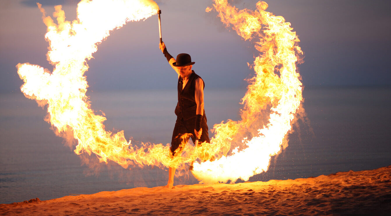 Firebeach.jpg: Performer wirbelt Feuer, kreisförmige Flammenspuren in der Abenddämmerung.