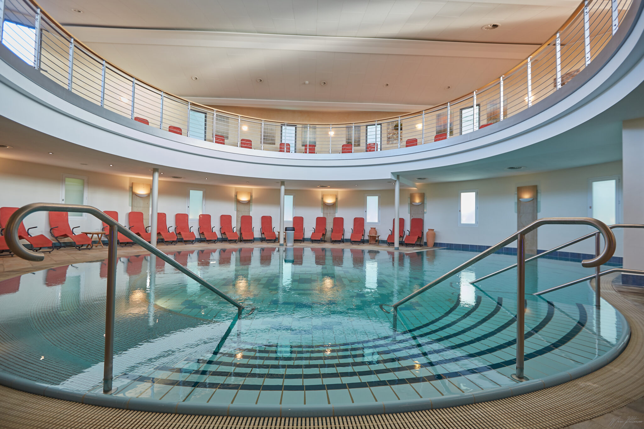 Meerwasser_schwimmbad.jpg: Rundes Hallenbad, rote Stühle, moderner Balkon darüber.