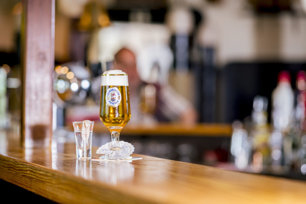 Damp_bar_scene.jpg: Bier und Schnapsglas auf einer lebhaften Holzbar.