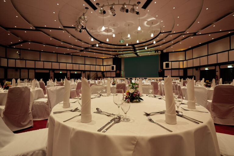 Familienhotel_silvester_bankett: Eleganter Bankettsaal, runde Tische, Bühne, weiße Dekoration.