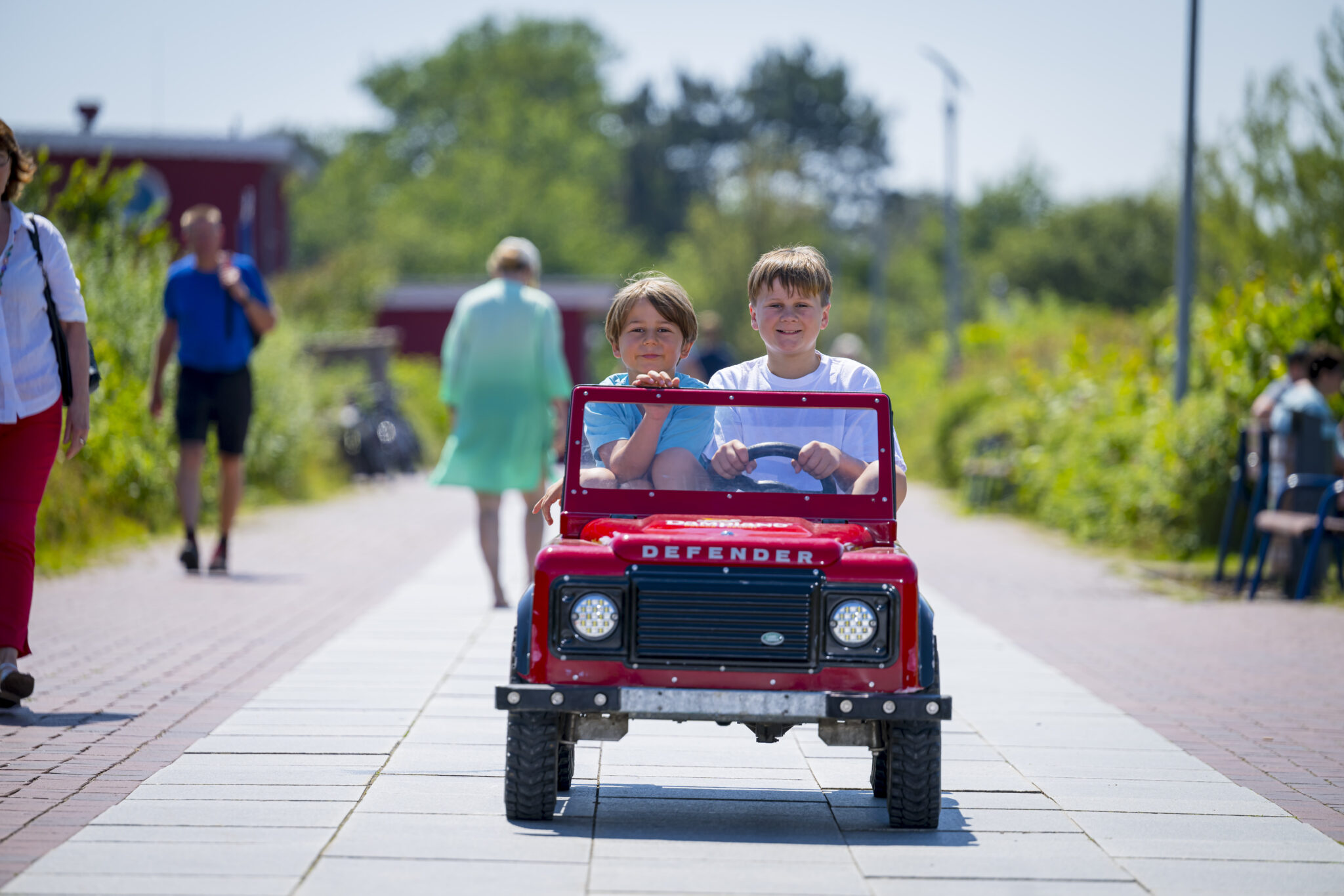 Defender-toy-car-children.jpg: Zwei Kinder im roten "Defender"-Spielzeugauto im Freien.