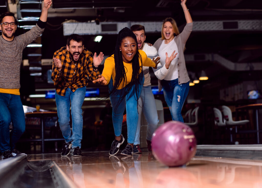 Bowling_group.jpg zeigt fünf Freunde, die jubeln, während einer eine lila Kugel kegelt.