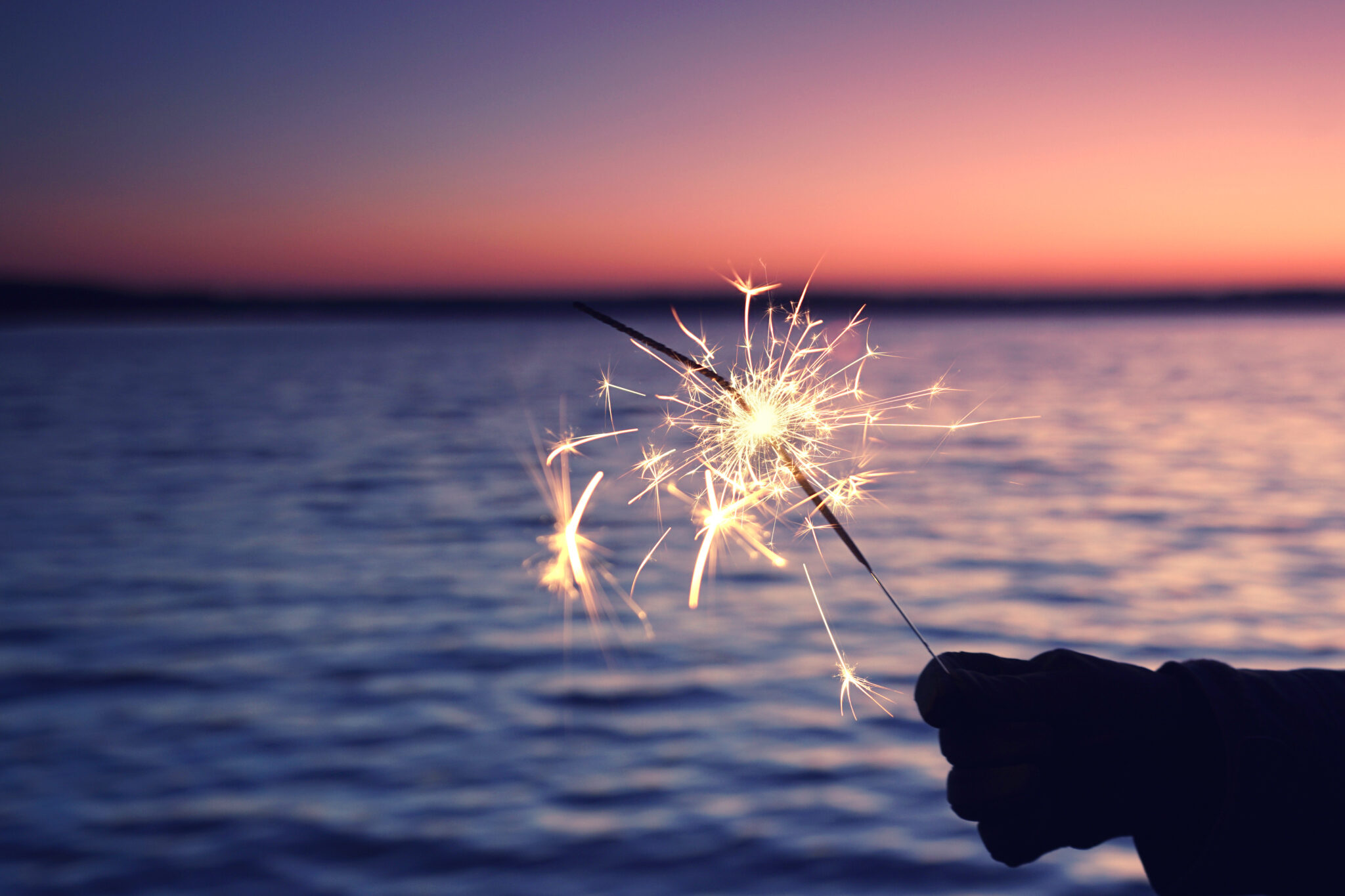 Sparkler_img12: Hand hält Wunderkerze bei Sonnenuntergang am See, bunter Himmel.