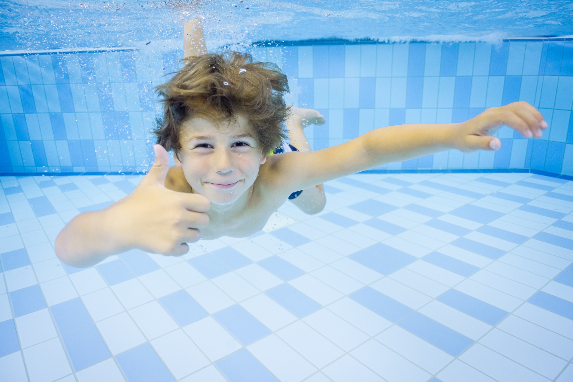 Winter-Wasserfreude_Entdeckerbad: Lächelndes Kind schwimmt unter Wasser, Daumen hoch, blaue Fliesen.
