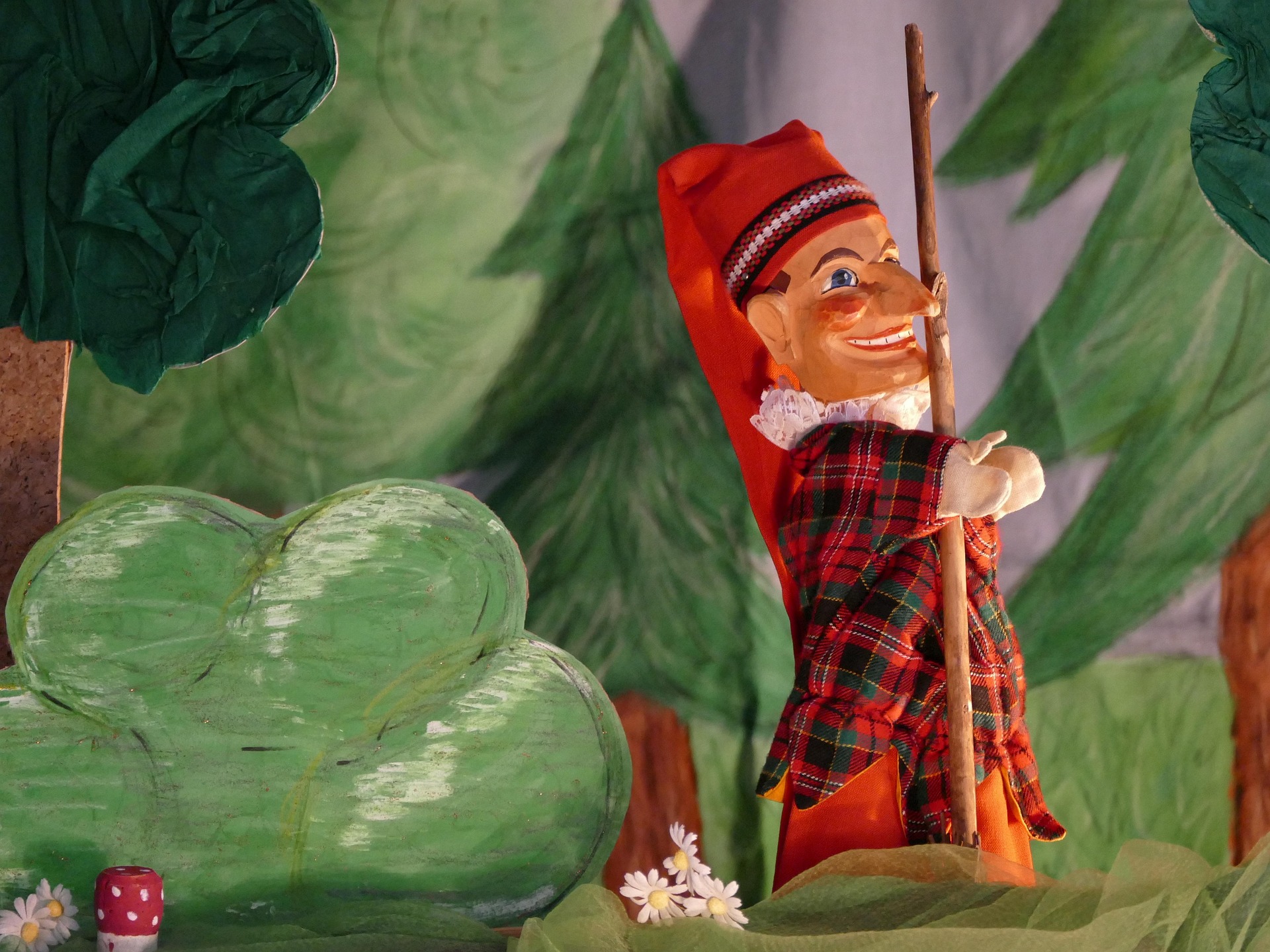 Wintermaerchen_mit_Kasper_Hoppe.jpg: Puppe mit rotem Hut inmitten von Bäumen, Blumen und Pilzen.