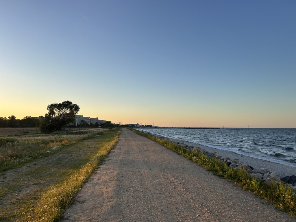 Lauf_zwischen_den_meeren_2026: Schotterweg am Meer, sonnenbeschienen, Gras und Felsen.