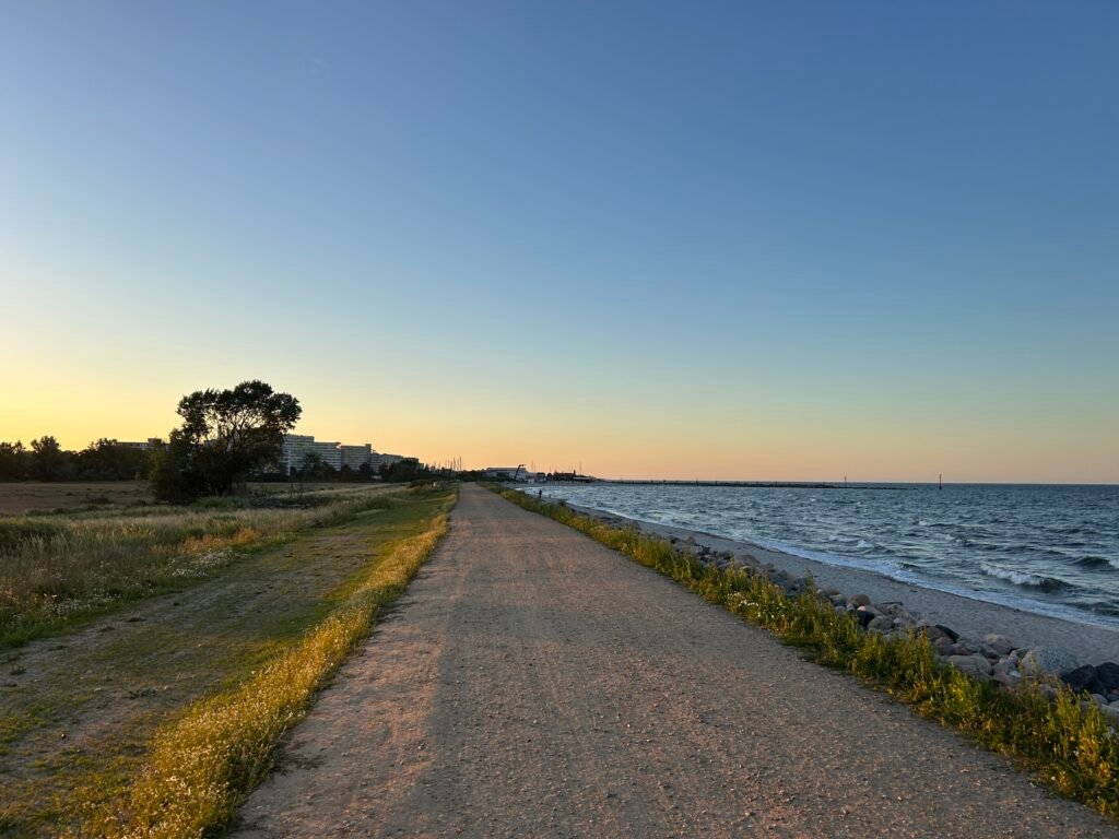 Lauf_zwischen_den_meeren_2026: Schotterweg am Meer, sonnenbeschienen, Gras und Felsen.