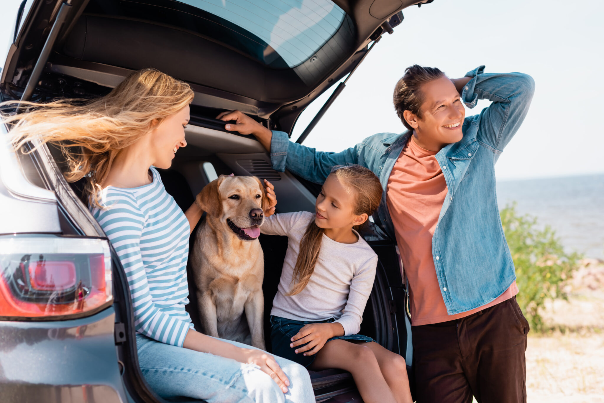 Familie lächelt am Kofferraum eines Autos, mit Hund.
