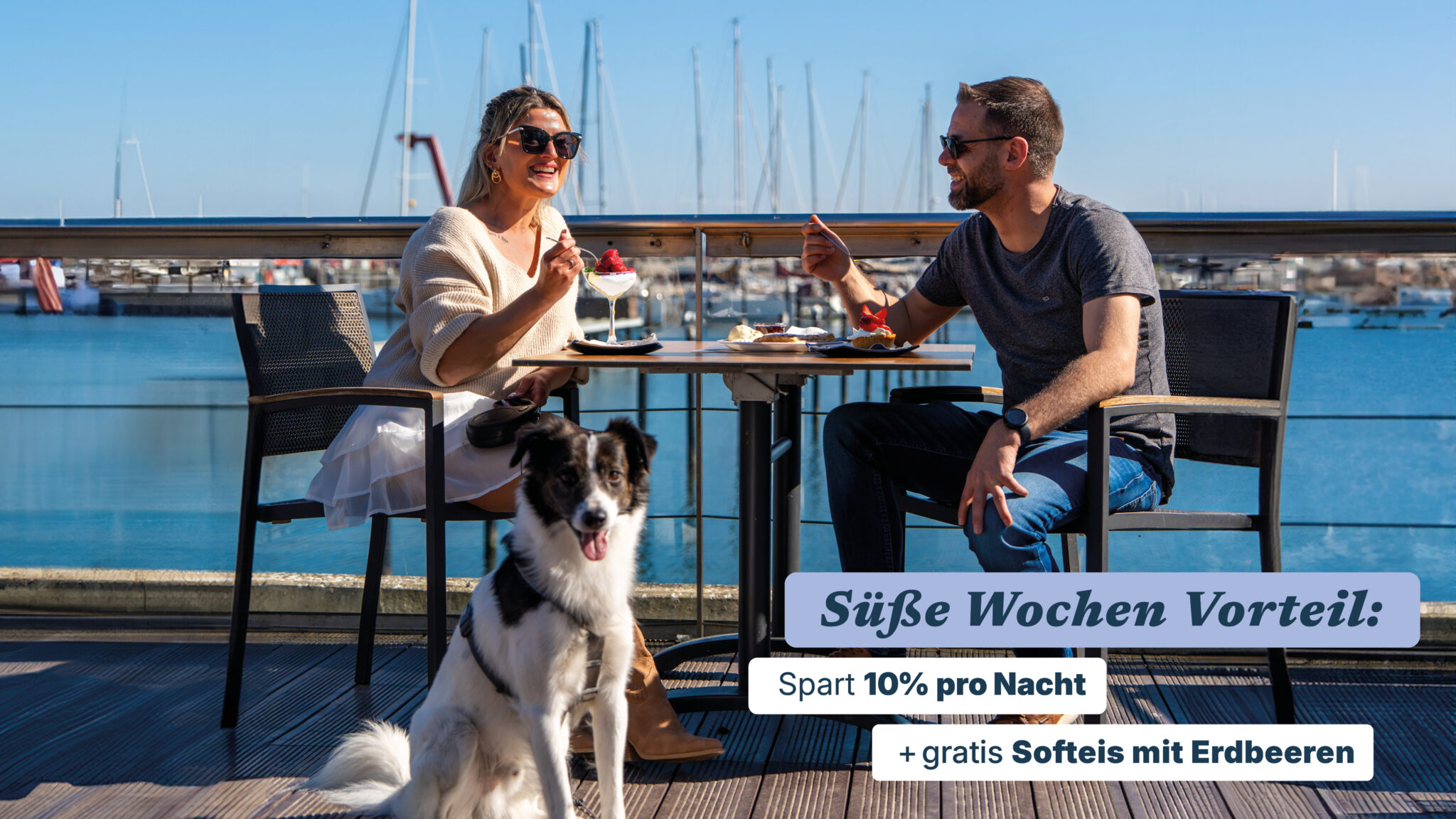 Eine blondhaarige Frau und ein braunhaariger Mann sitzen auf der Terrasse eines Restaurants und essen Erdbeereis. Ein Schwarz-weißer Hund sitzt davor.
