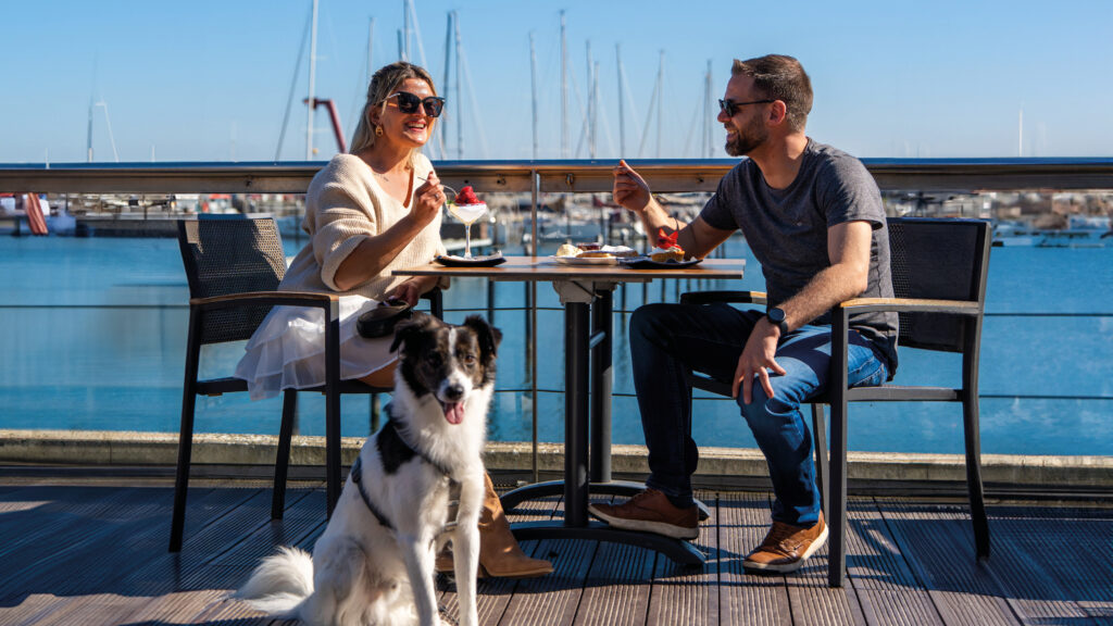 Eine blondhaarige Frau und ein braunhaariger Mann sitzen auf der Terrasse eines Restaurants und essen Erdbeereis. Ein Schwarz-weißer Hund sitzt davor.