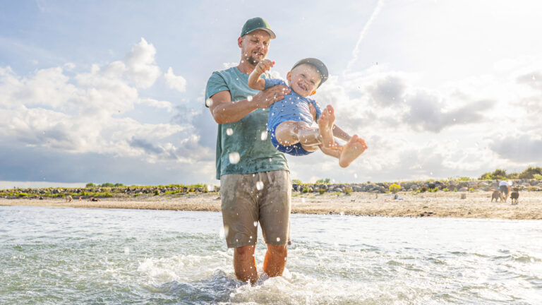 Parent_child_beach_fun.jpg: Lächelndes Kind, das über spritzendes Wasser schwingt, Ostsee Resort. Fotograf: Oliver Franke