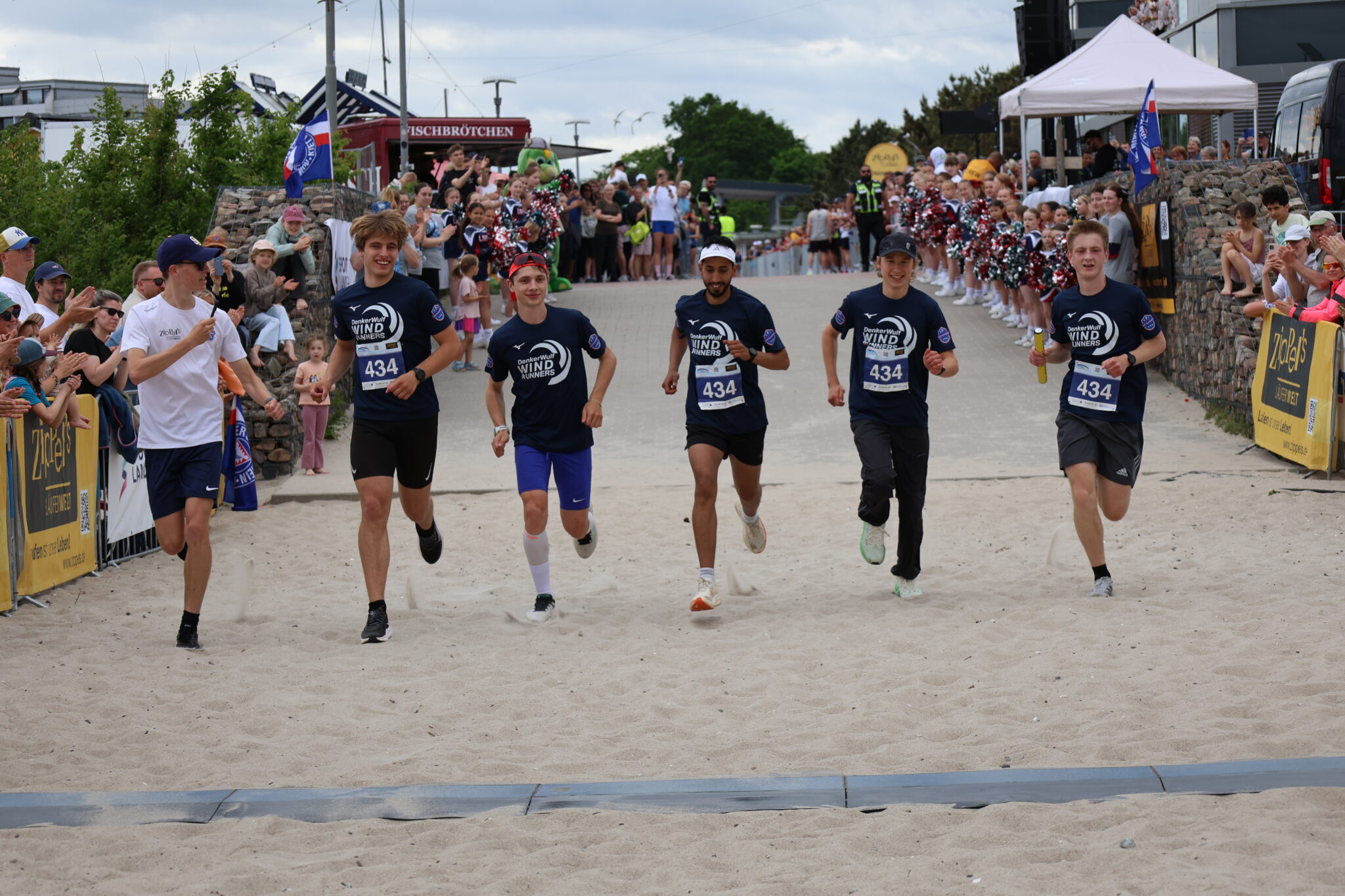 Finishline_runners.jpg: Fünf Läufer in der Nähe des Ziels; die Menge jubelt, Transparente winken.