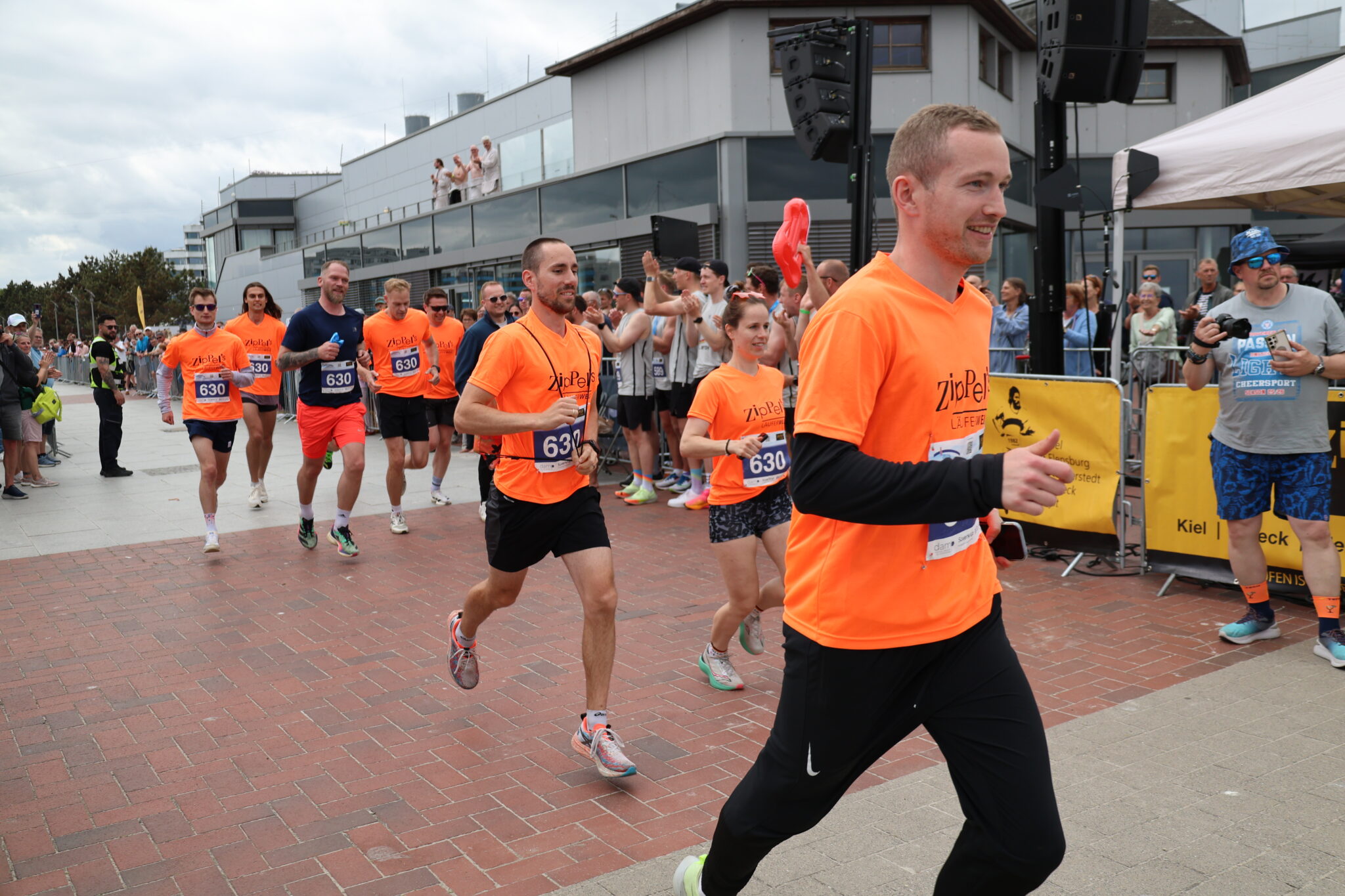 Raceevent5.jpg zeigt Läufer in Orange, jubelnde Menschenmengen neben den Gebäuden.