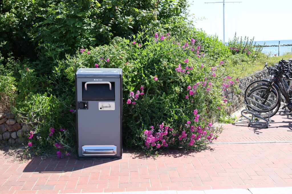 Nachhaltig_bin.jpg: Graue Mülltonne neben einem gepflasterten Weg, Blumen, Sträucher, Fahrräder.