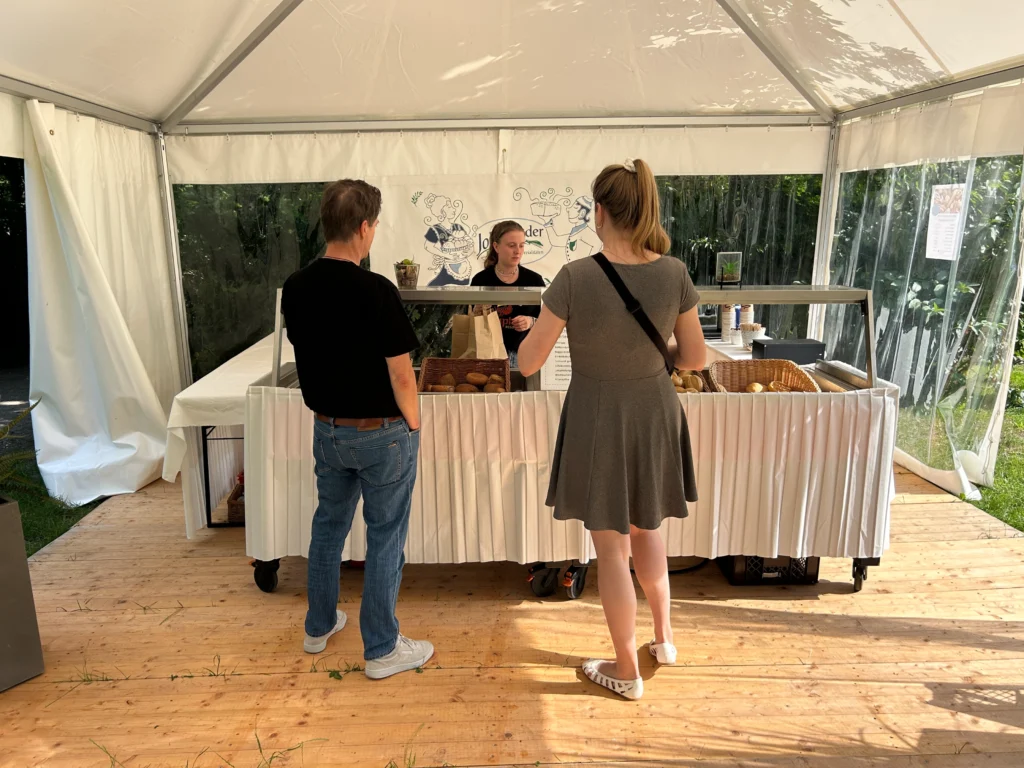 Ostsee_damp_bakery.jpg: Zwei Personen kaufen Brot an einem Brötchenverkaufsstand im Freien.