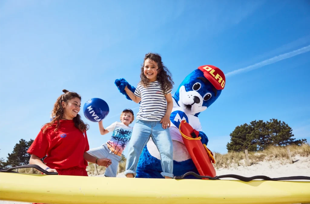 Strandfest-damp: Frau, Kinder, blaues Robben-Maskottchen in einem gelben Kanu, Strand.