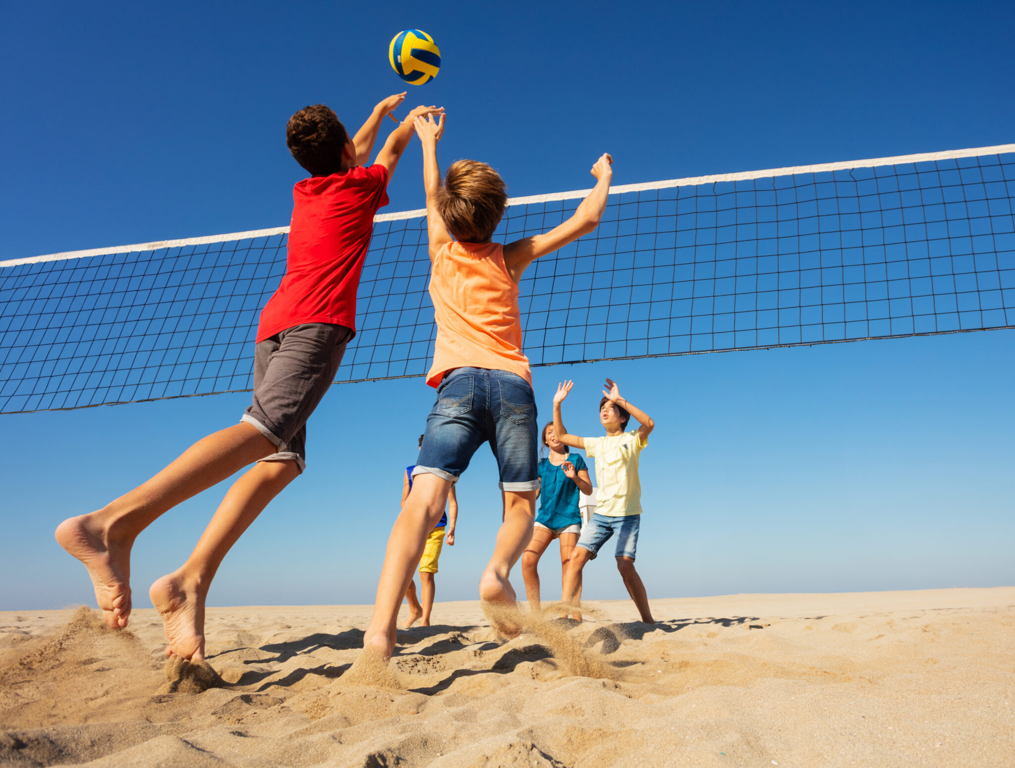 6kids_beach_volleyball: Kinder spielen Volleyball am Sandstrand, in Sommerkleidung.