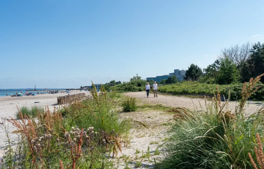 Ostsee_resort_damp: Menschen spazieren auf einem sandigen Strandweg, blauer Himmel, Gebäude in der Ferne.