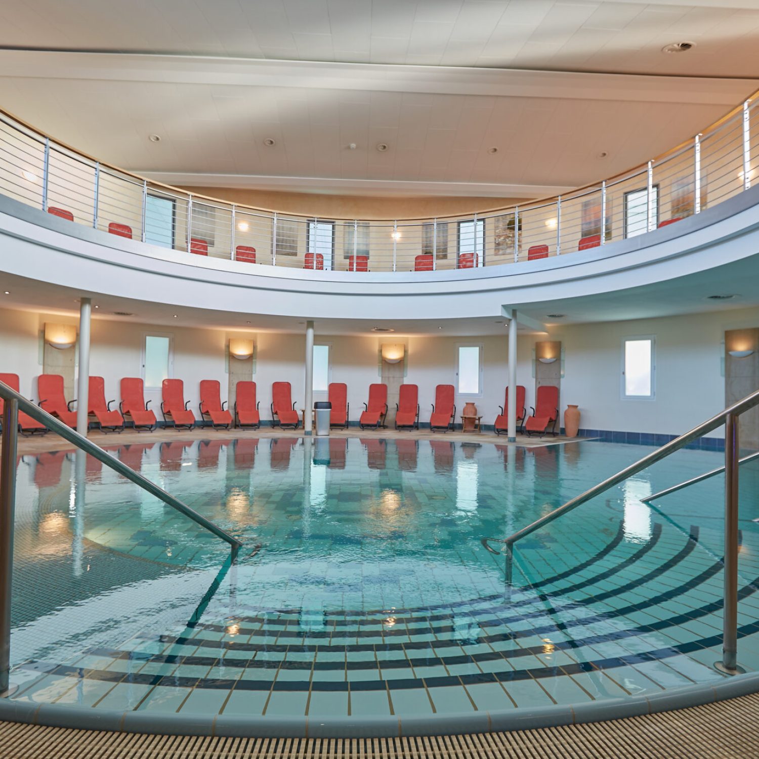 Meerwasser_schwimmbad.jpg: Rundes Hallenbad, rote Stühle, moderner Balkon darüber.
