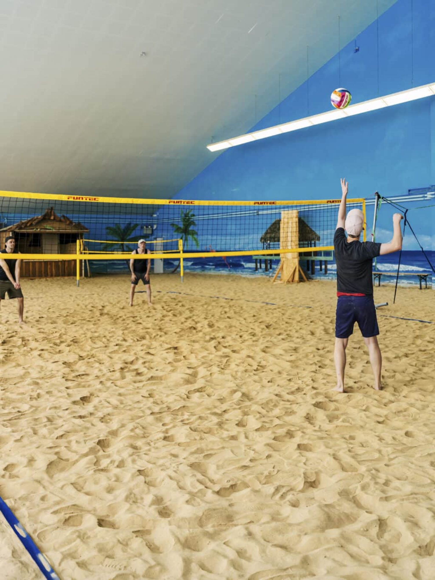 Drei spielen drinnen Beachvolleyball, Wandmalerei aus Palmen und helle Lichter.
