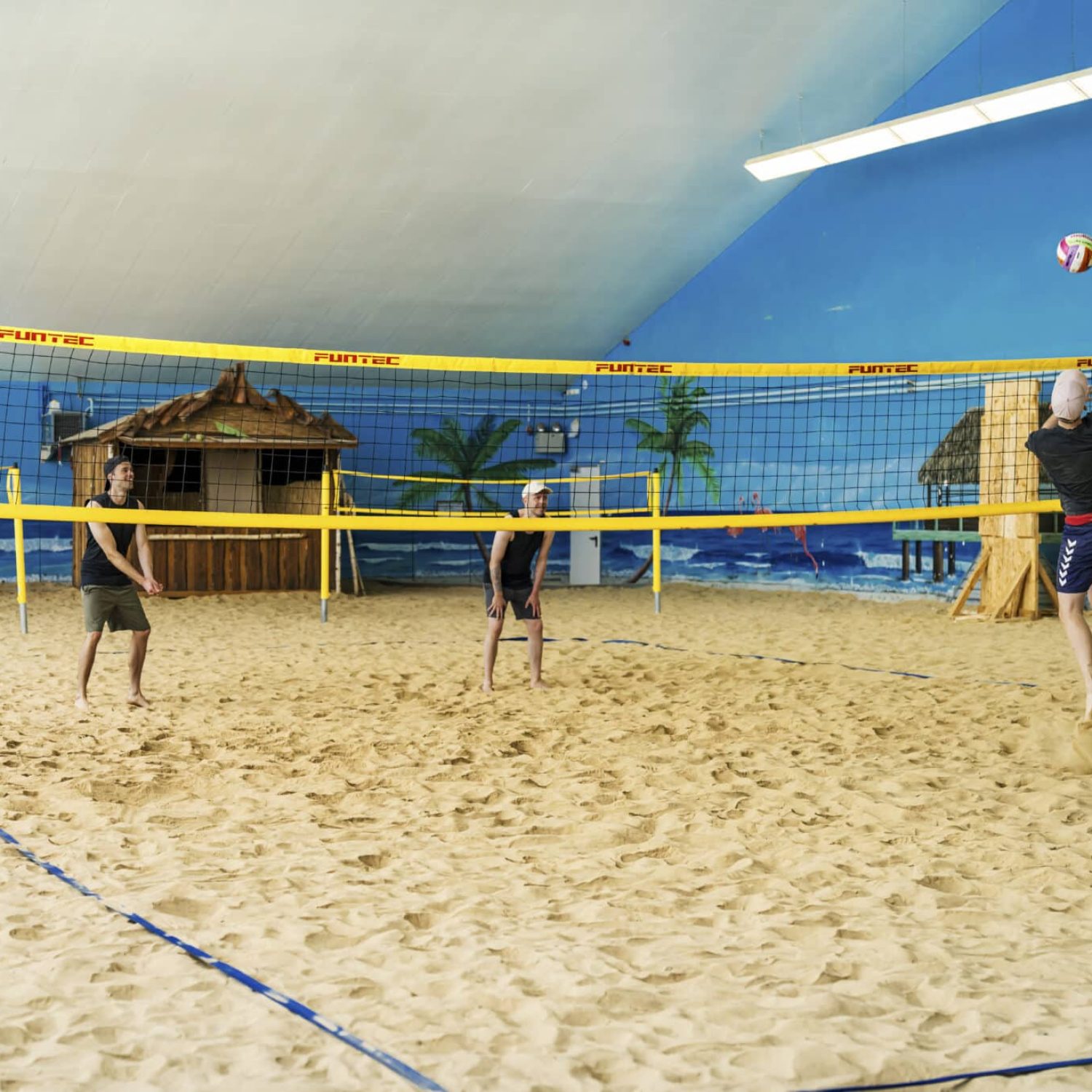 Drei spielen Indoor-Beachvolleyball auf Sand mit tropischem Dekor.