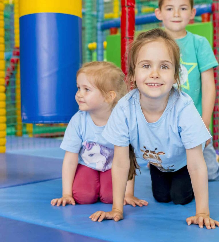 Drei lächelnde Kinder spielen zusammen auf Matten in einem Indoor-Spielplatz.