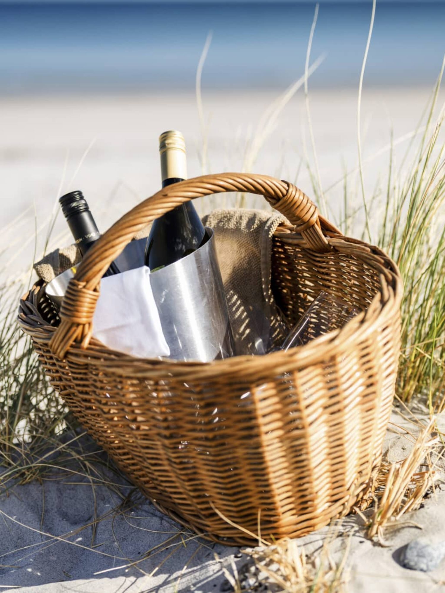 Weidenkorb mit Weinflaschen auf Sand, das Meer dahinter verschwommen.