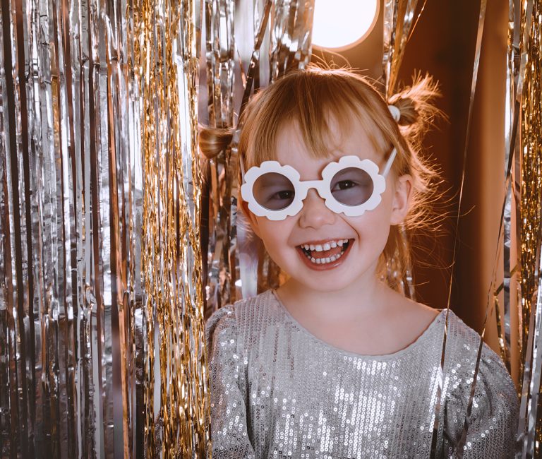 Silvester_familienhotel_child.jpg: Lächelndes Kind mit Brille, Paillettenoberteil, Lametta-Kulisse.