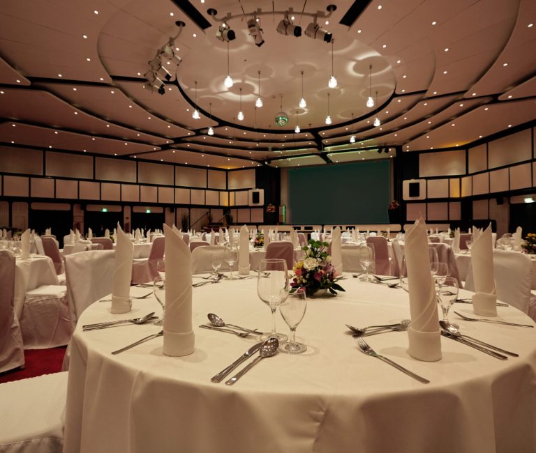 Familienhotel_silvester_bankett: Eleganter Bankettsaal, runde Tische, Bühne, weiße Dekoration.