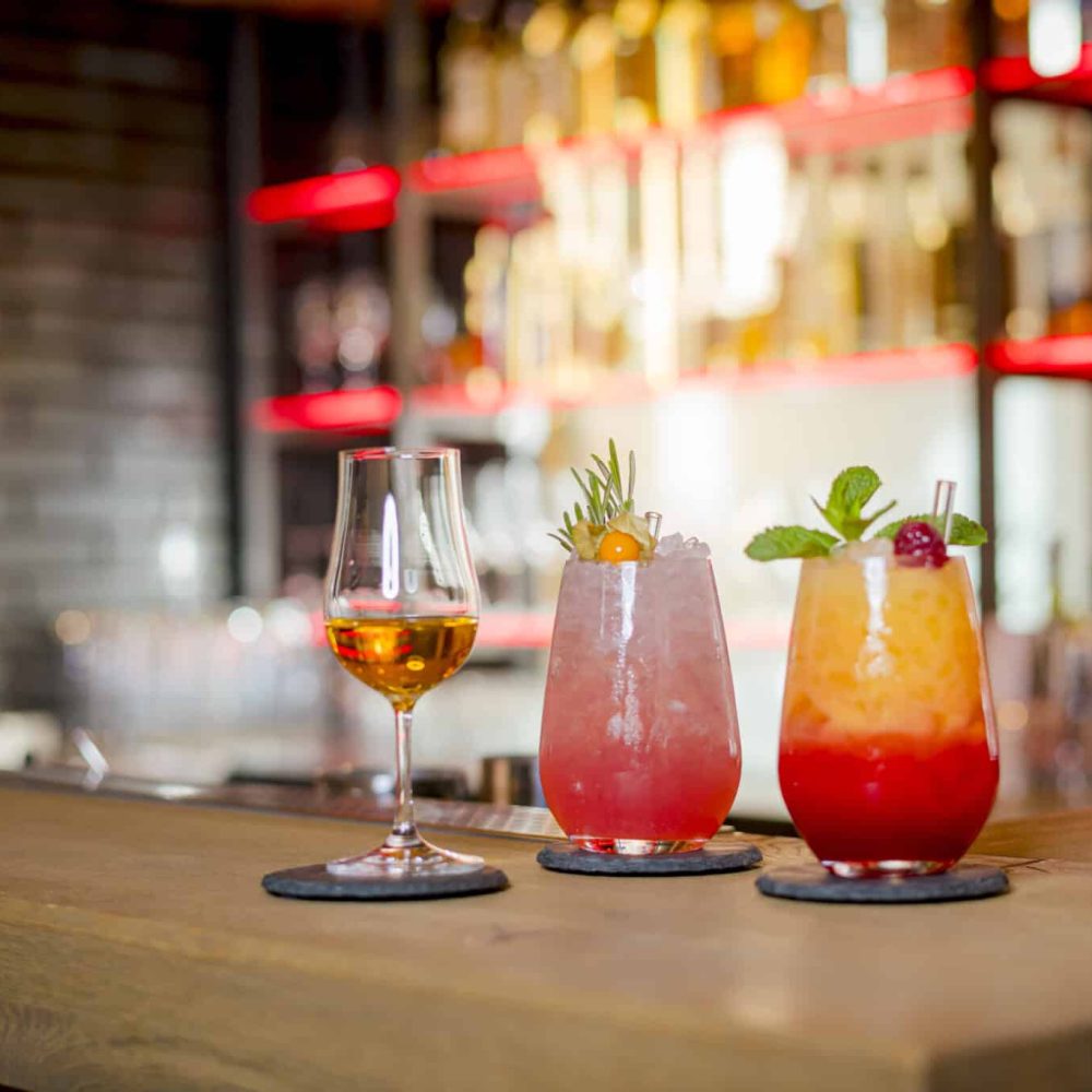 Bernsteinfarbener Schnaps und bunte Cocktails auf Untersetzern an der lebhaften Bar.